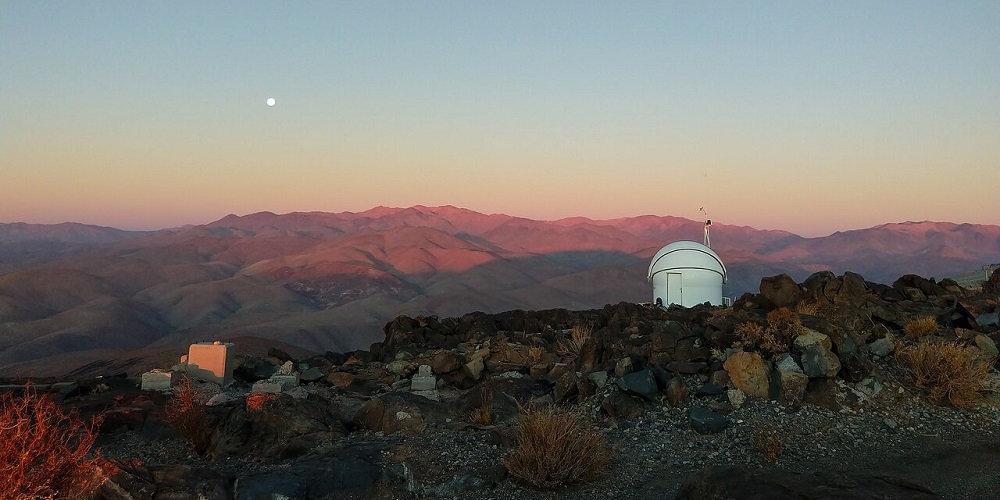 De Test-Bed Telescope 2, gestationeerd op de ESO-sterrenwacht op La Silla in Chili, kijkt bij zonsondergang uit over de roze getinte Atacama-woestijn. Links zien we de maan opkomen.